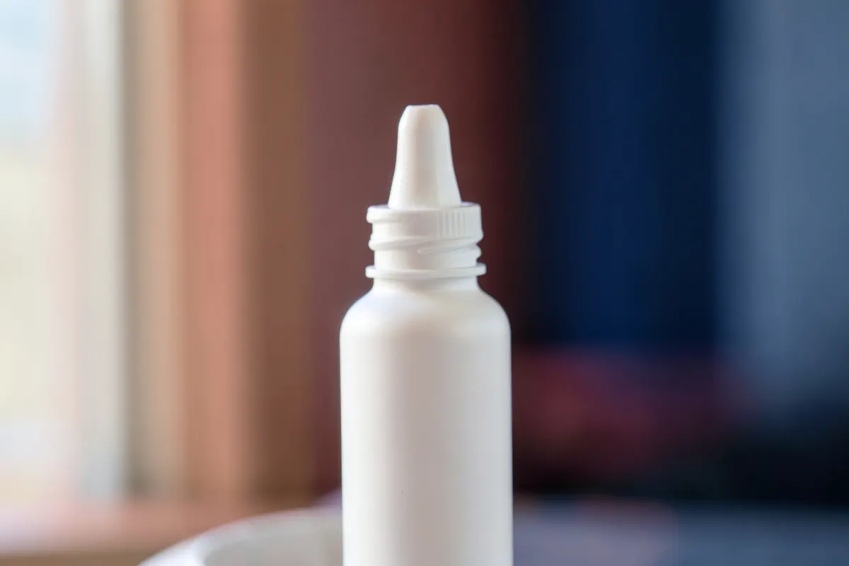 Mão segurando um frasco de spray nasal com um fundo desfocado de um ambiente interno.